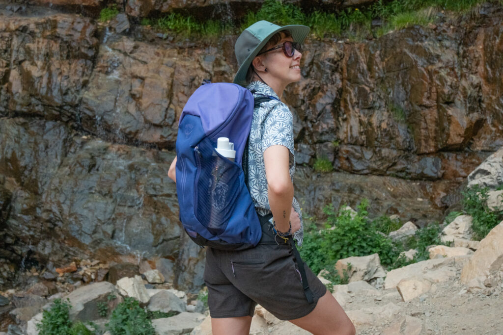 A woman on a trail wears a purple Osprey Hikelite LT 16 daypack.