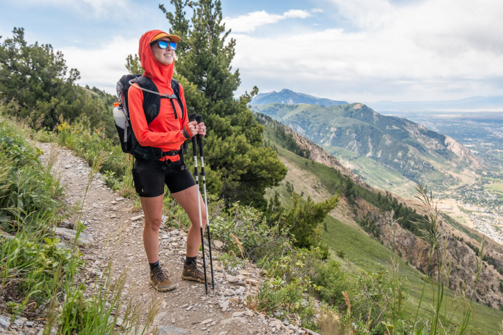 A woman stands on a hiking trail with trekking poles and an ultralight Rab Syclon 28 backpack.