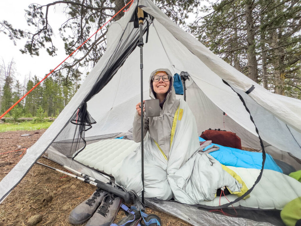 A woman sits in a Hyperlite unbound tent wrapped in a sleeping bag and holding a mug. It's grey and rainy.