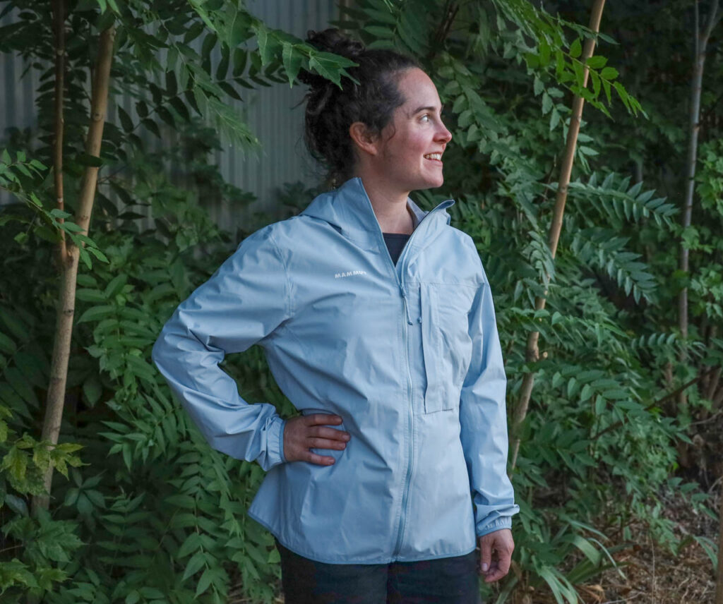A woman stands under trees in a Mammut Duncan Light HS hooded ultralight rain jacket.