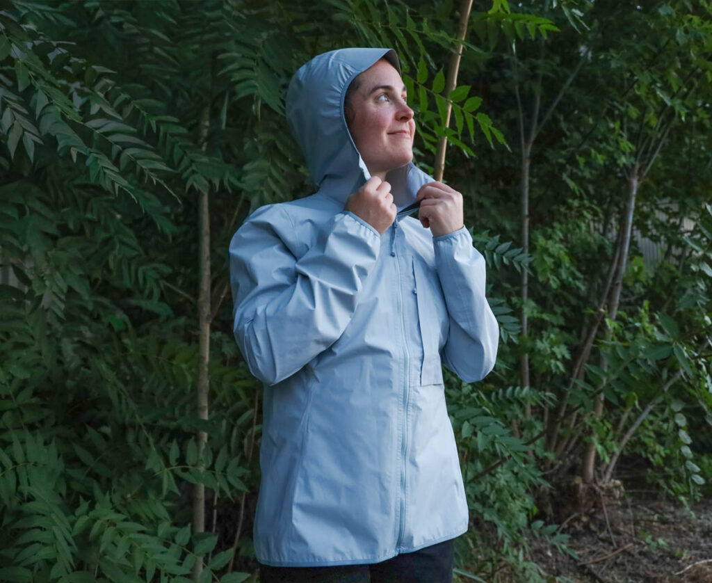 A woman stands under trees in a Mammut Duncan Light HS hooded ultralight rain jacket (hood up).