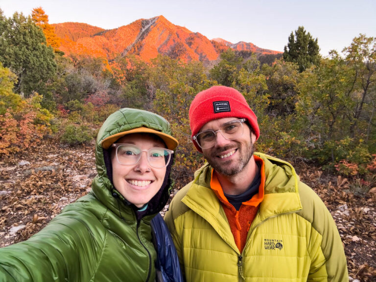 A man and woman in jackets smile from an autumn peak.